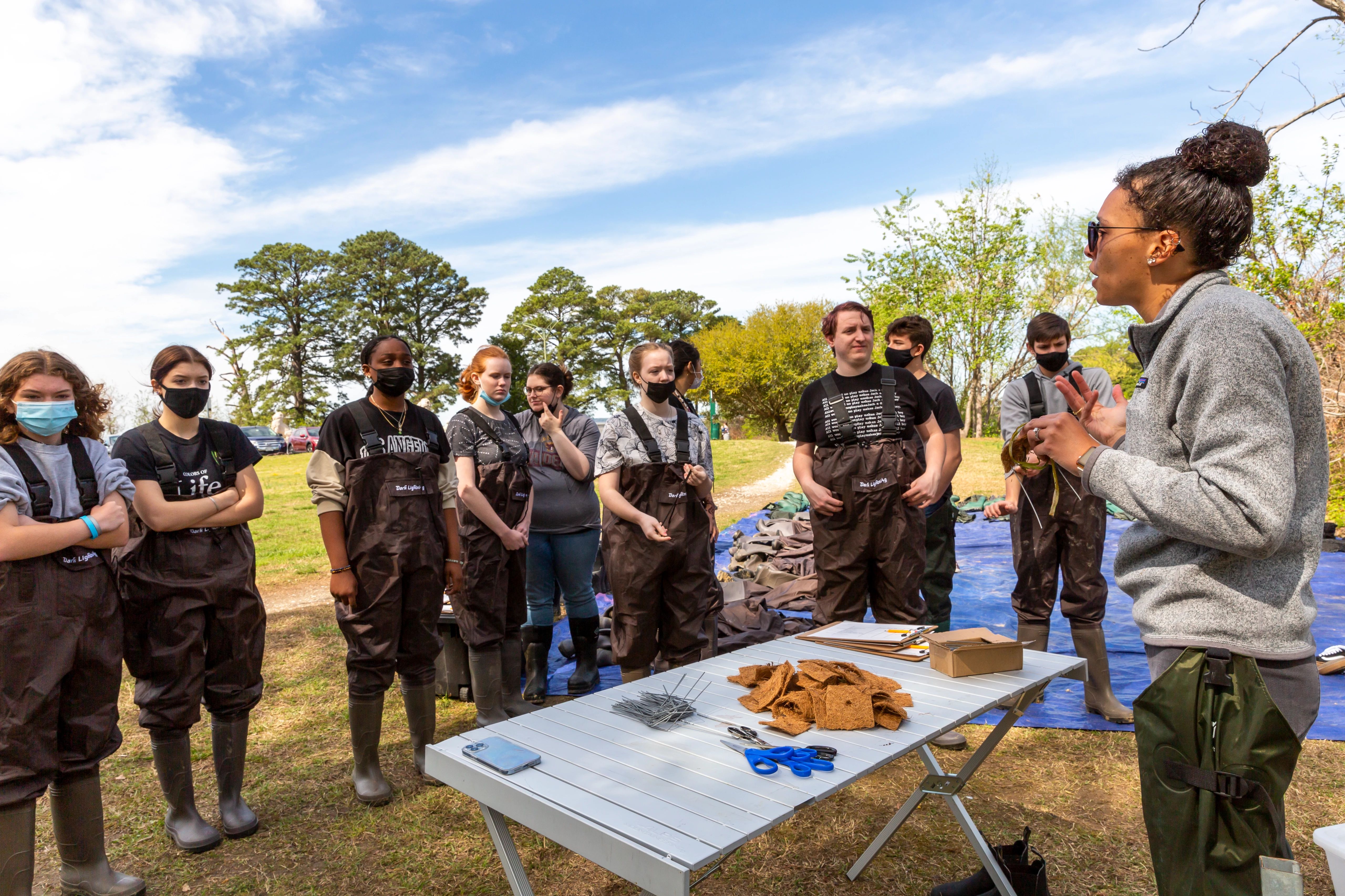 Science Educator Shantelle Landry is outside in The Mariners' Park along with a dozen high school students from Newport News. Shantelle is explaining their next steps with the students watching her and wearing overall waders and boots. They are preparing to get into Mariners' Lake to do water quality testing.