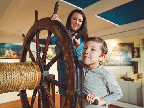 Mother and child turn a large ship's wheel. 