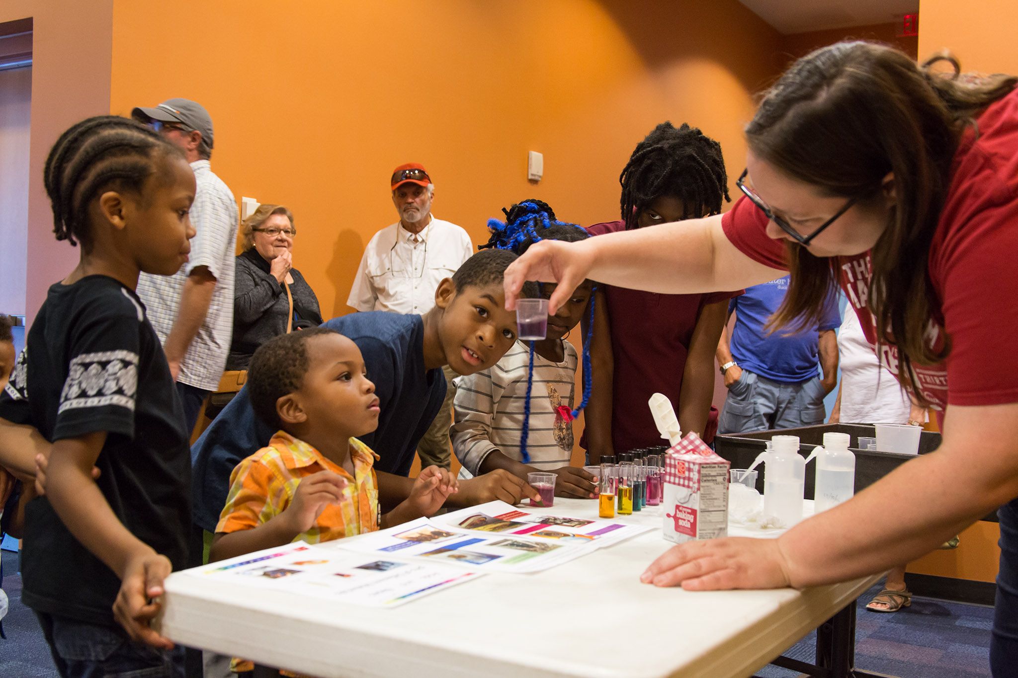 Research Scientist Molly McGath is seen showing young black children a scientific experiment using water colors and small beakers. The children are peering into the glass Molly is holding in front of them.