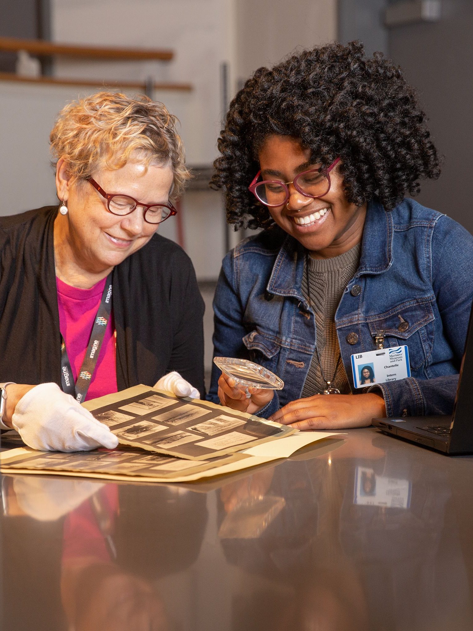 Mariners' Library team member sitting at table with a student volunteer. They are both looking at prints and the student is holding a magnifying glass to see the images in detail. 