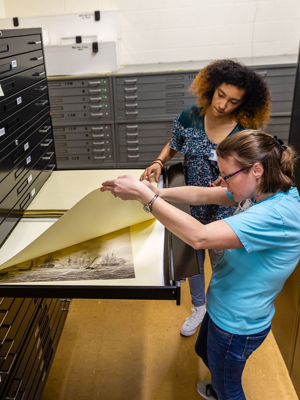 A white and black woman looking through a large flat file draw of works on paper.