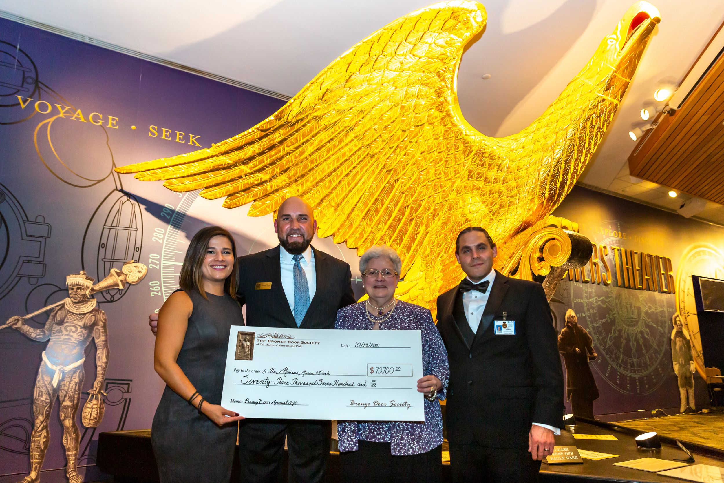Bronze Door Society Member Cynthia Katz presents a check to Mariners' team members in front of the golden Lancaster eagle figurehead.