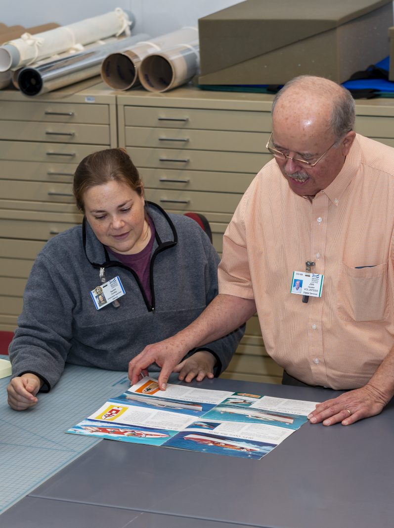 Young white female and older white male at a work table looking at a Chris-Craft marketing brochure. In the background are large flat files and rolled plans.