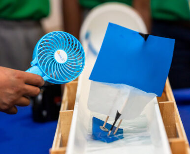 A handmade small boat has its ability to float tested in shallow waters inside a cut-out PVC pipe. Someone is holding a hand held fan near the small boat's sails.