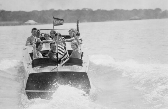 A black and white photo of a group of white females and males riding away for the viewer in a Chris-Craft boat.