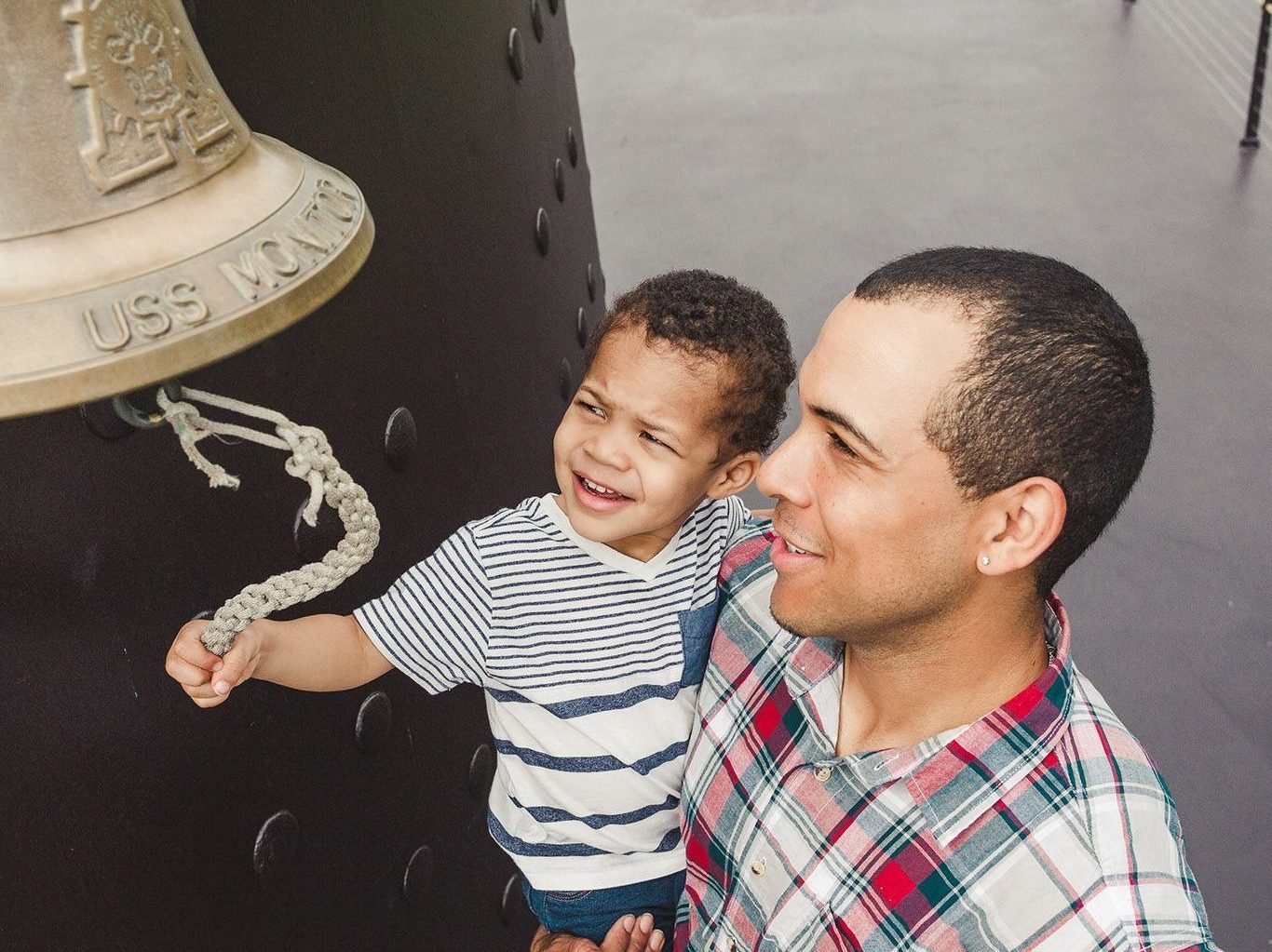 Father holding toddler son who is ringing the bell on the USS Monitor replica.