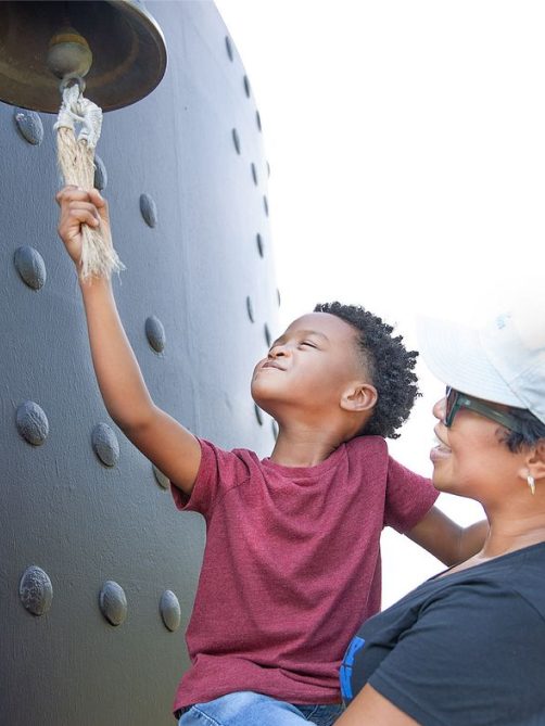 Little boy ringing the bell on the USS Monitor replica, while an adult female holds him up.