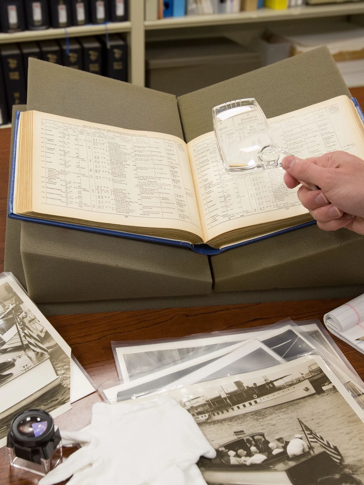 Color photo of a log book open on a table. A white persons hand is holding a magnifying glass. Black and white photos are spread out under the book. There is also a white glove and magnifying loop on the table. In the background is a bookshelf with books on it.