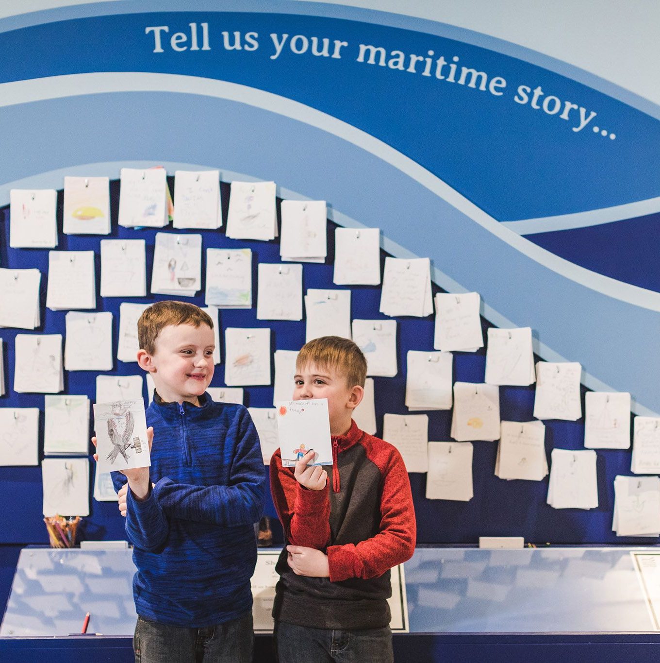 two children hold up their drawings while standing in front of story wall.