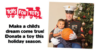 Toys for Tots advertisement. The photo shows a member of the United States Marine Corps with a young child, posing in front of a Christmas tree. The Marine is wearing the Blue Dress uniform. Both the Marine and the child are holding a basketball together. Next to this image is the Toys for Tots train logo, with text underneath that says, "Make a child's dream come true! Donate a toy this holiday season."