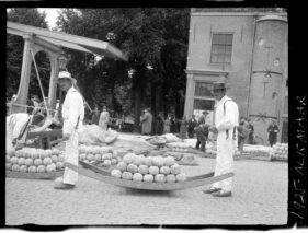 Men, dressed in traditional attire, carry a curve-shaped barrel with balls of cheese stacked on top.