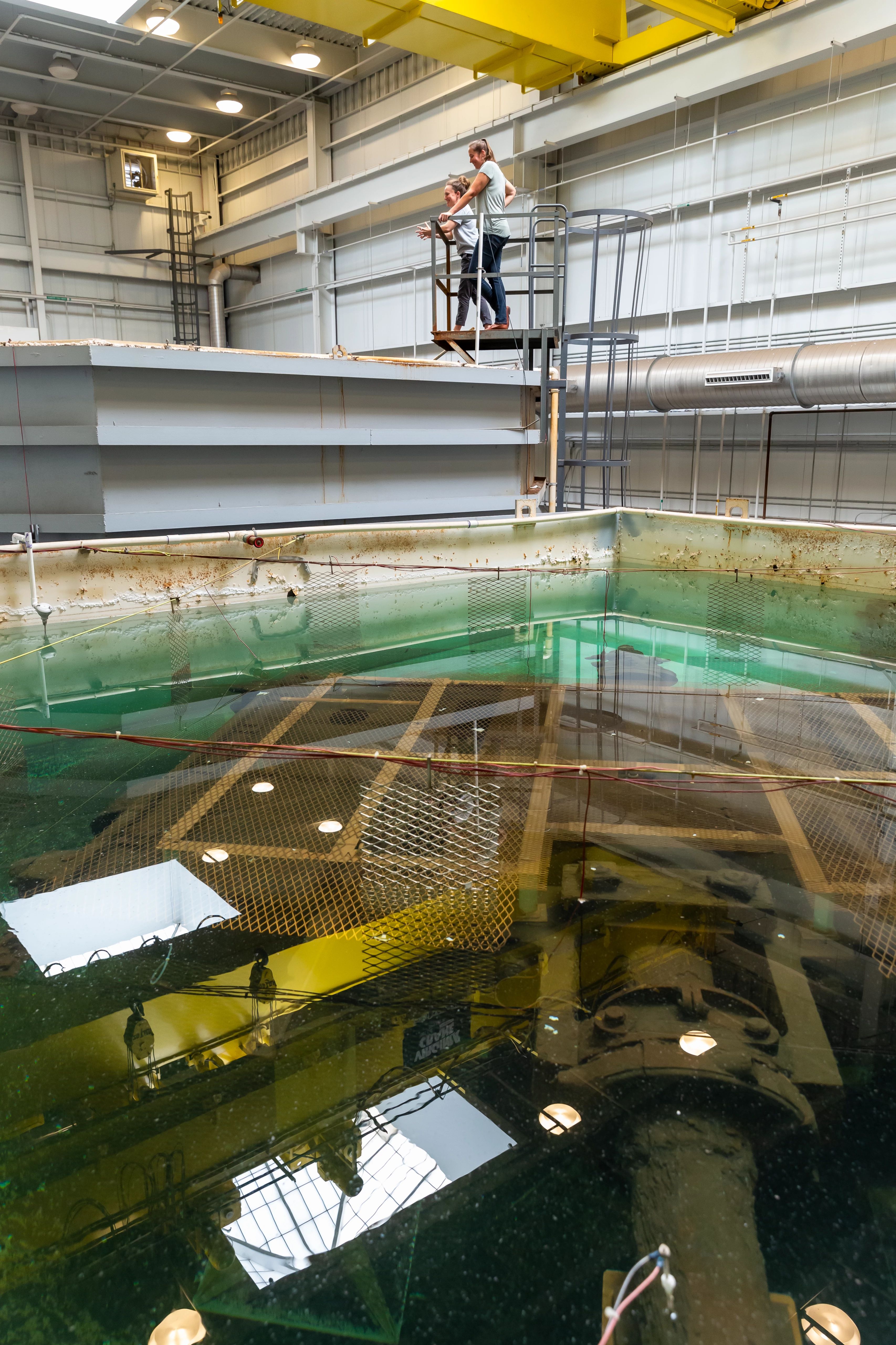 Image of large, industrial tanks holding corroded artifacts submerged in liquid. Two conservators stand on a platform over the larger tank.