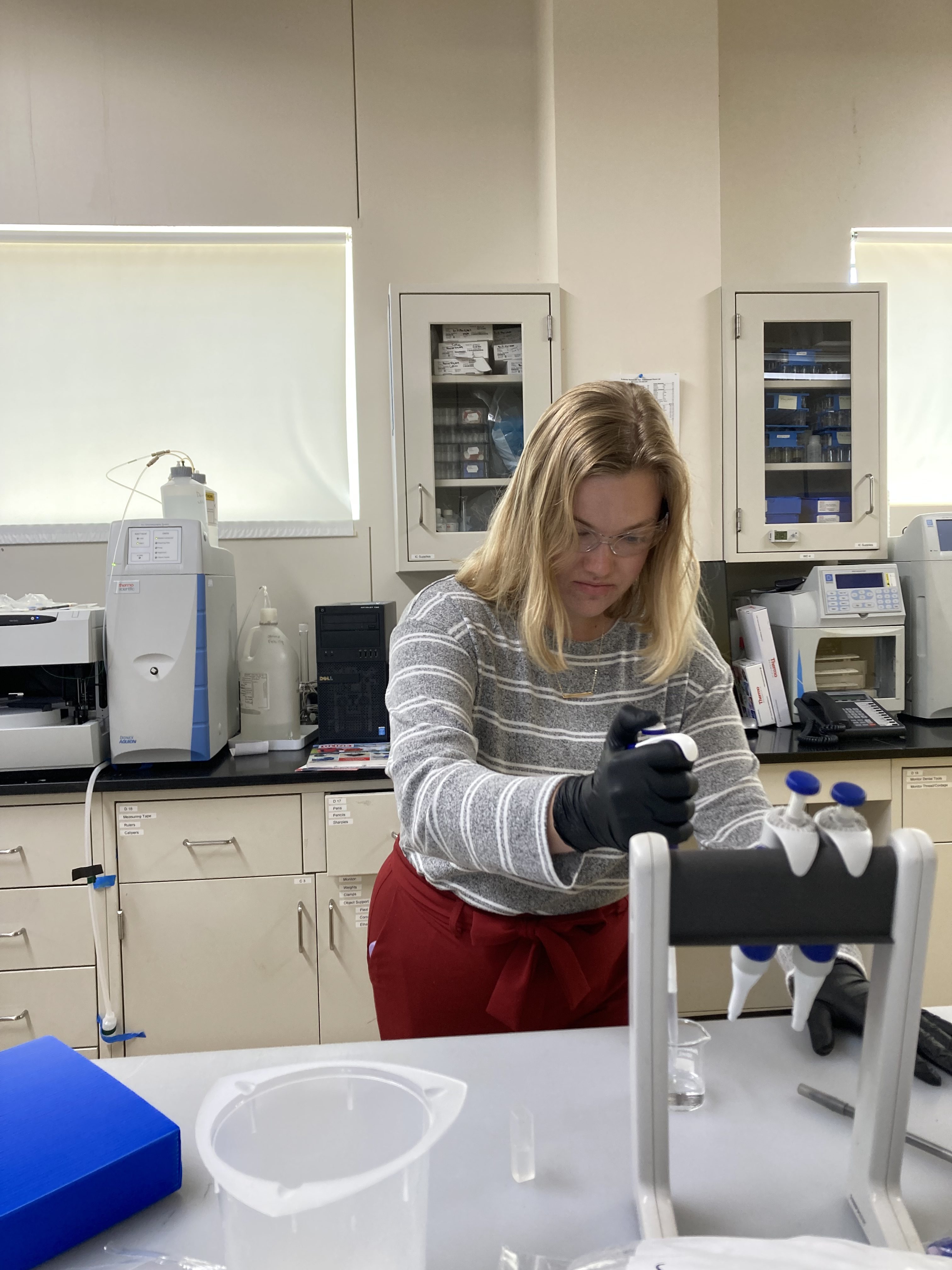 Image of scientist in a lab wearing safety goggles and gloves, and holding a device to transfer small amounts of clear liquid from beaker to smaller container.