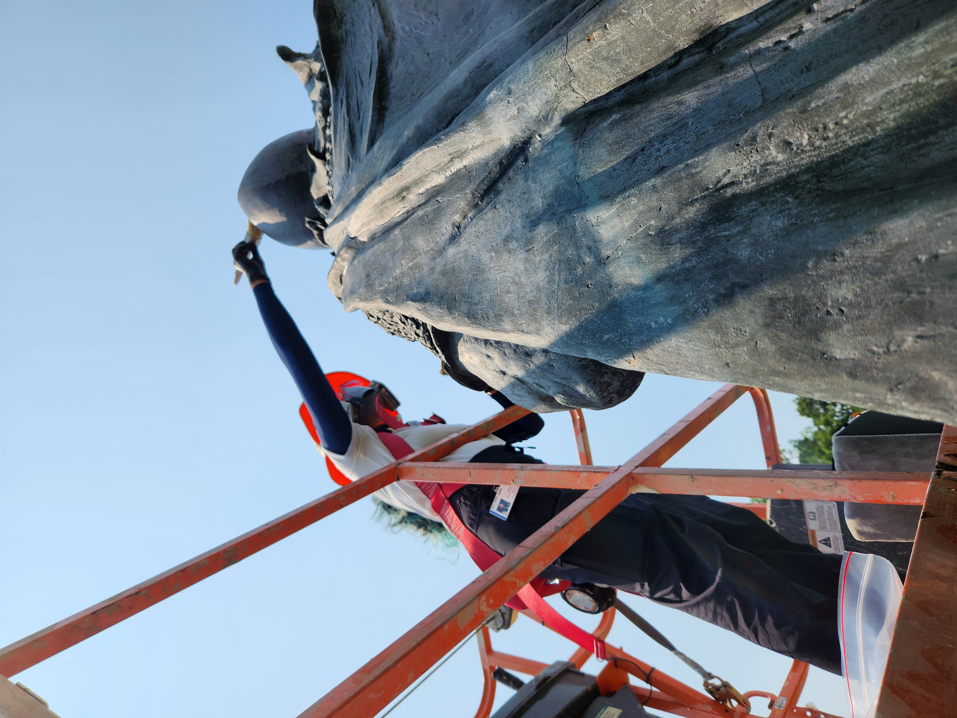 Image of Conservation intern Marimar Bracero Rodríguez on an orange lift applying wax with a natural brush to the head of Leifr Eiriksson, Son of Iceland.