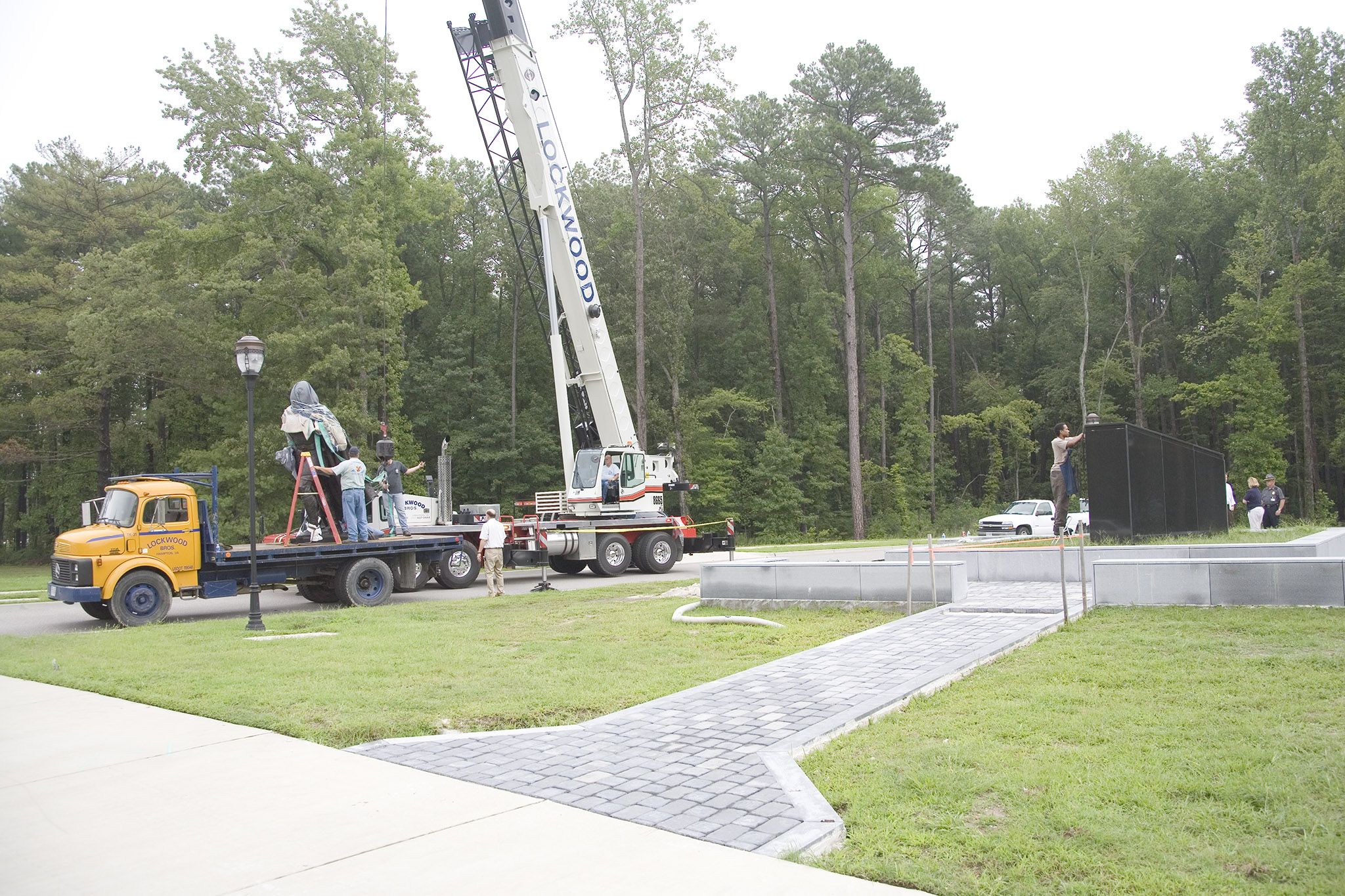 Image of a flatbed truck with the statue on it and crane trailing the truck near the footer block where the statue was to be mounted.