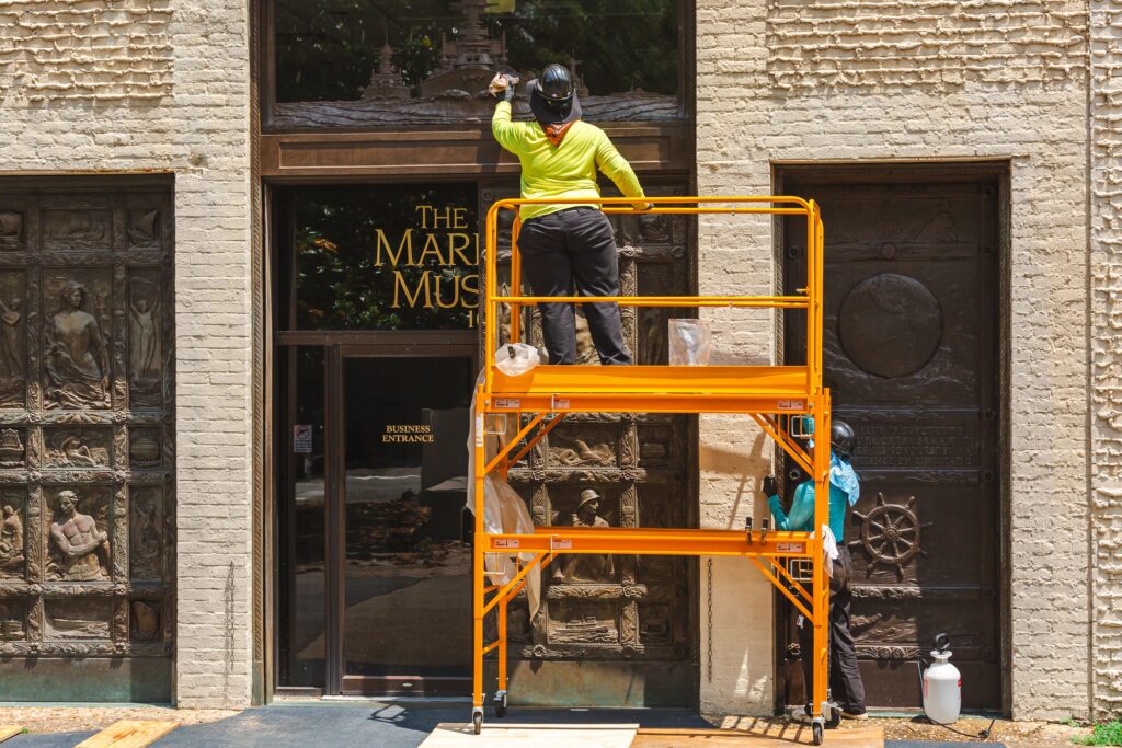 Image of Senior Objects Conservator Erik Farrell on a scaffold cleaning the top panel and Intern Marimar cleaning a door.