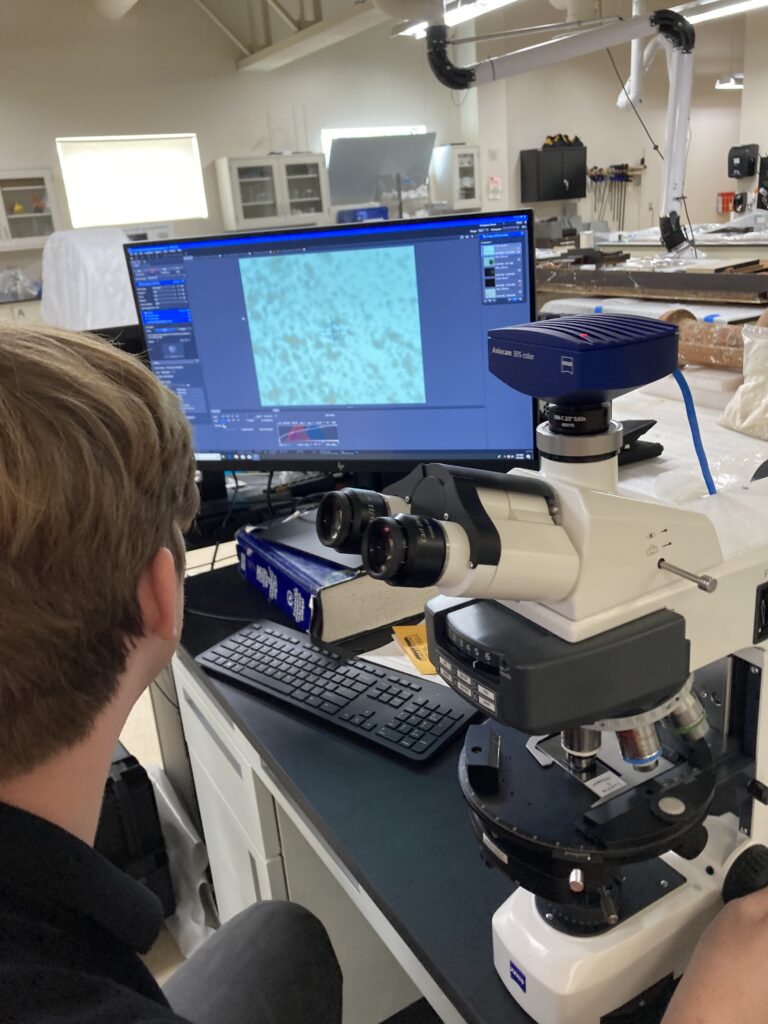 Image of Harrison Biggs using a microscope, focusing on a sample. The microscope image is projected on a computer screen in the background.