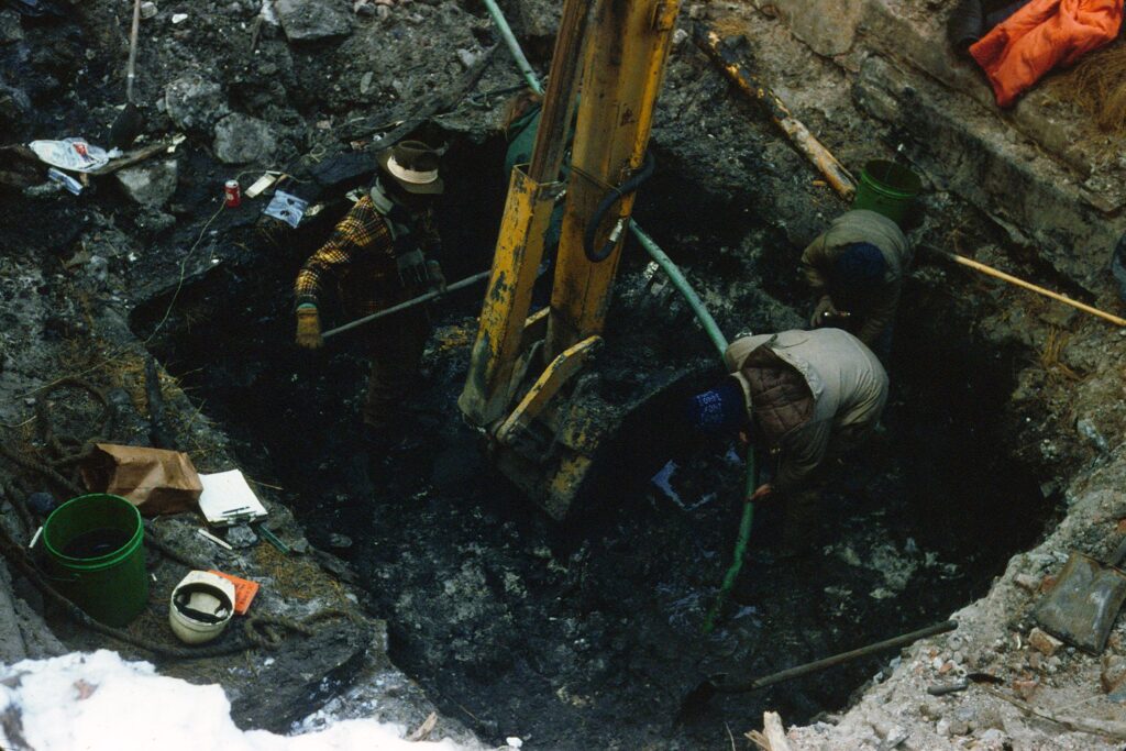 Historical photo of two workers assisting an excavator in a shallow rectangular hole, surrounded by equipment and debris, including a bucket and hard hat.