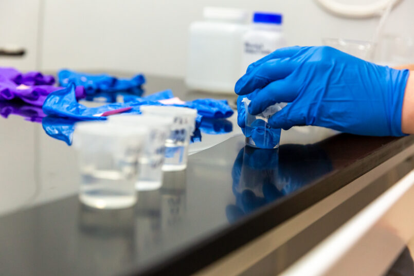 Close up image of water samples in the fume hood. A single finger from a glove sits submerged in water in each of three beakers. Conservation intern Harrison Biggs works to prepare a new sample in the background.