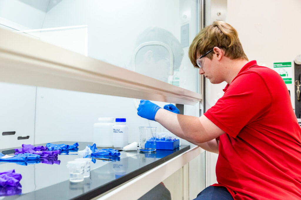 Image of Conservation intern Harrison Biggs working at the fume hood, removing the forefinger from a glove and placing it in a small beaker filled with water.