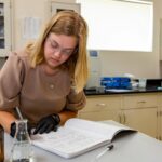 Photo of scientist writing in laboratory notebook with chemical jars and flask in foreground.