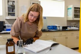 Photo of scientist writing in laboratory notebook with chemical jars and flask in foreground.