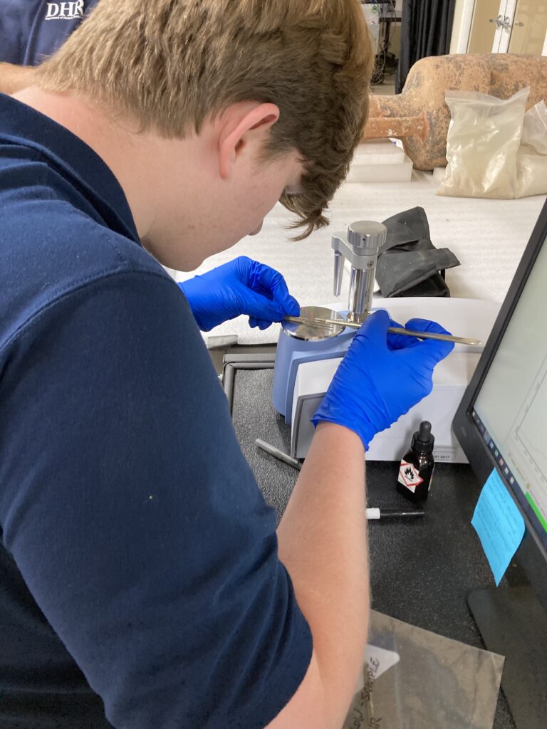 Image of Conservation intern Harrison Biggs working with the ATR-FTIR. He is using a scalpel to scrape some material off of a sample onto the ATR-FTIR.