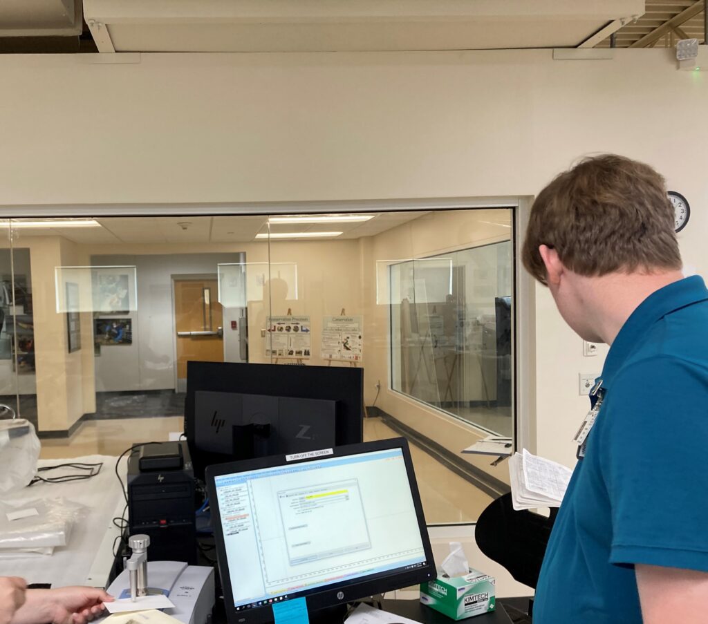 Image of Conservation intern Harrison Biggs working with the ATR-FTIR. He operates the software as Lindsey secures the object in the instrument.