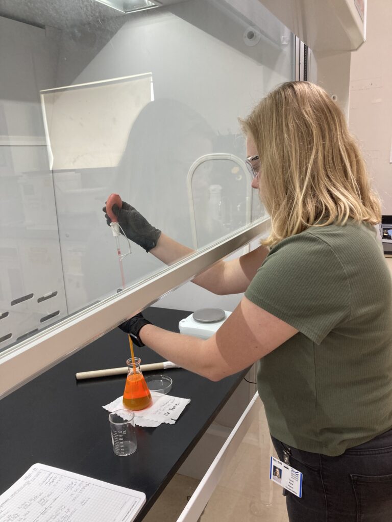 Photo of scientist working in a fume hood to transfer bright orange liquid.