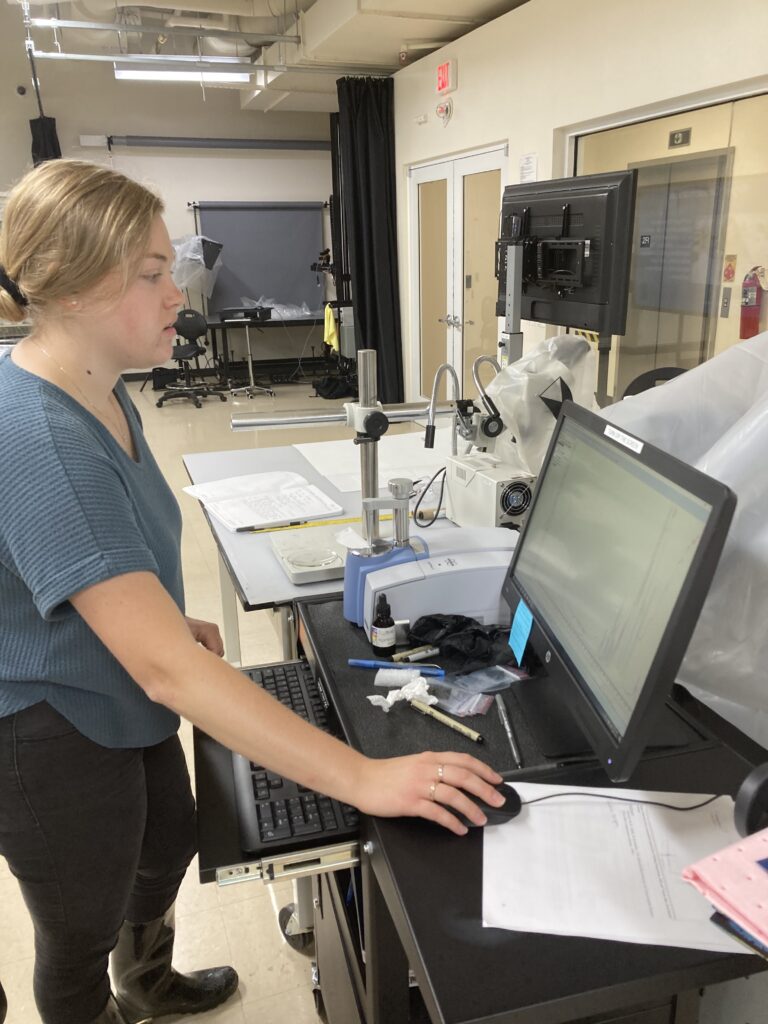 Photo of scientist standing at computer displaying spectrum, next to instrument holding small piece of paper.