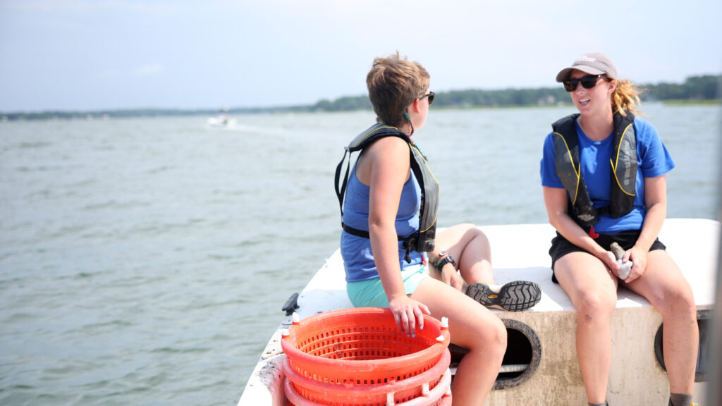 Julie Leucke (L) and Jess Lutzow (R) sit on the bow of the Chesapeake Bay Foundation’s skiff as they head out to plant oysters on restoration reefs in the Lynnhaven River.