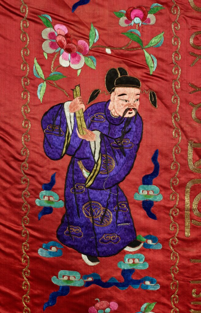 On the tapestry, a figure of a god dressed in purple holds a pair of cylindrical tablets.