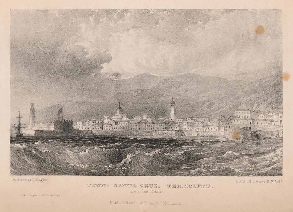 An image of a town with mountains in the background and water in the foreground.