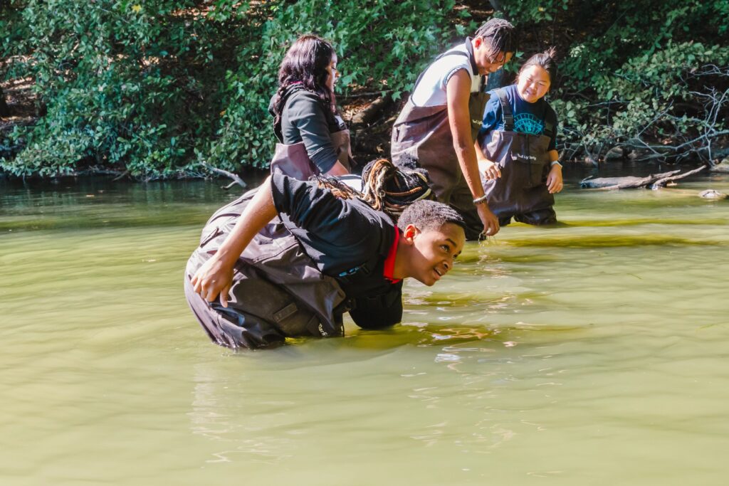 Children in waders stand in a lake surrounded by foliage.