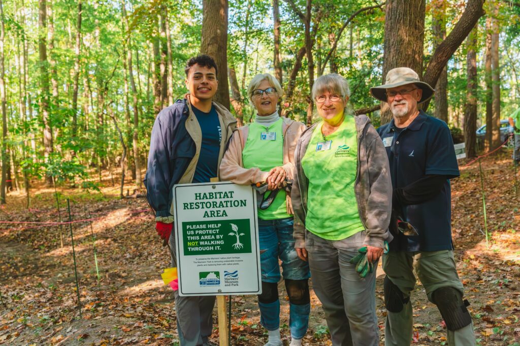Four people stand in a park behind a sign that reads "Habitat Restoration Area"