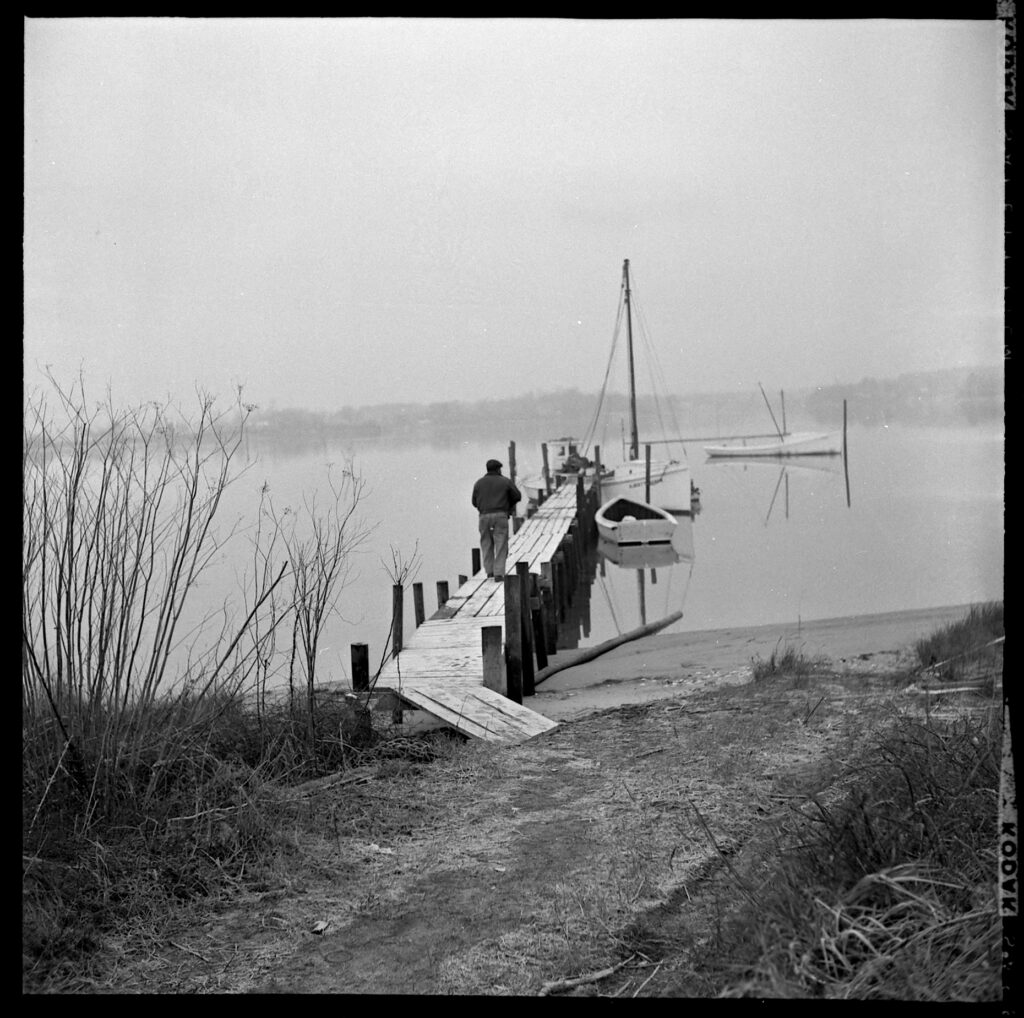 Black and white image of a man walking along a pier on a misty day with boats floating in the water.