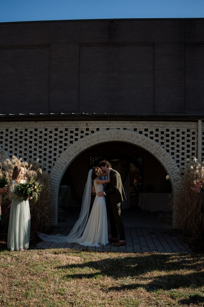 A bride and groom embrace. A bridesmaid holding a bouquet watches.