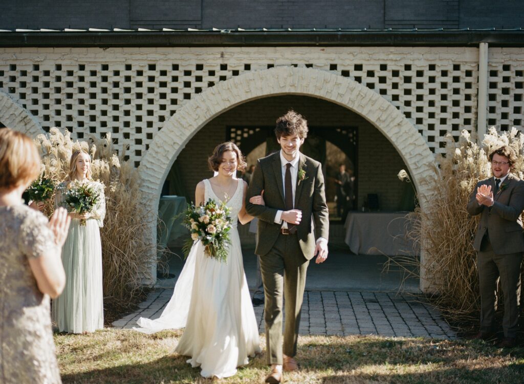 A bride and groom walk with arms linked as others applaud.
