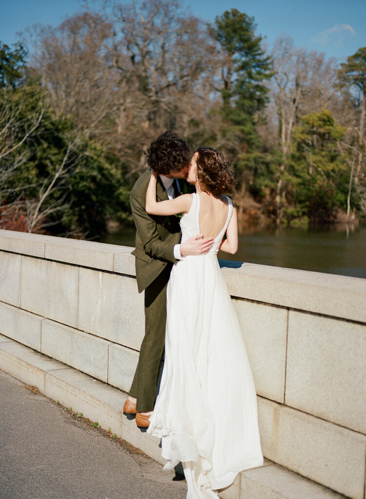 A bride and groom kiss with water and trees in the background.