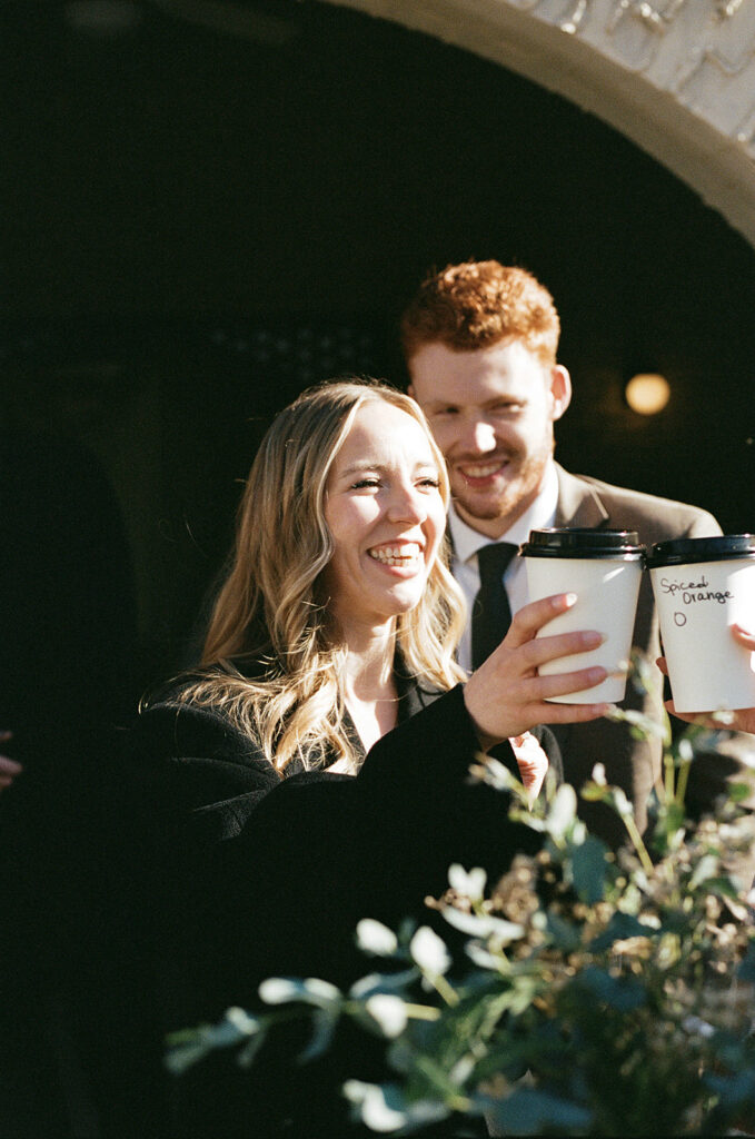 A man and woman smile. The woman is holding up a cup to another person's cup for a cheers.