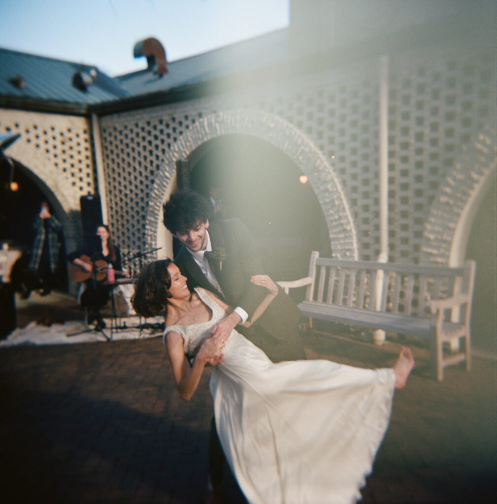 A groom dips a bride while dancing with a woman smiling and playing the guitar looking on.
