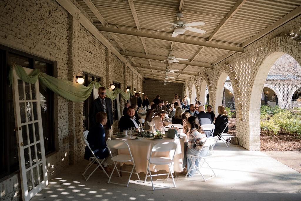 Sunlight fills a covered patio as several guests are spread out, chatting around round tables and chairs.