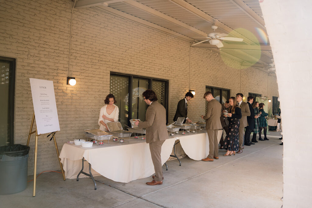 A sign on a stand that says "lunch" is at the end of row of tables with food on them that people are holding plates in front of.