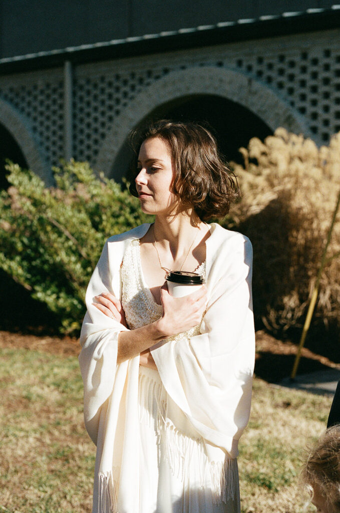 A bride stands in the sunlight holding a coffee cup.