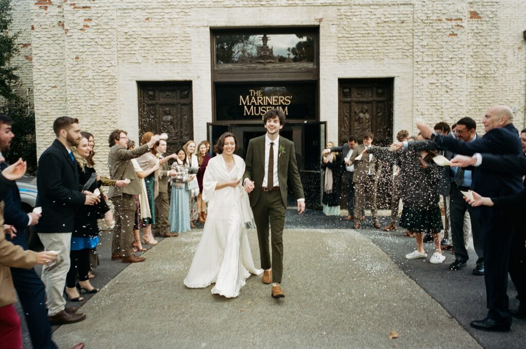 A bride and groom walk between 2 rows of people throwing a white substance. The couple is smiling. The building includes "The Mariners' Museum" on the enrance.