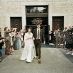 A bride and groom walk between 2 rows of people throwing a white substance. The couple is smiling. The building includes "The Mariners' Museum" on the enrance.
