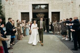 A bride and groom walk between 2 rows of people throwing a white substance. The couple is smiling. The building includes "The Mariners' Museum" on the enrance.