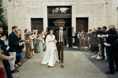 A bride and groom walk between 2 rows of people throwing a white substance. The couple is smiling. The building includes "The Mariners' Museum" on the enrance.