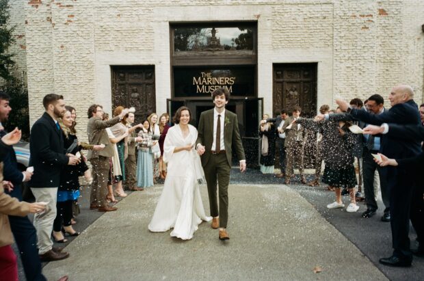 A bride and groom walk between 2 rows of people throwing a white substance. The couple is smiling. The building includes "The Mariners' Museum" on the enrance.