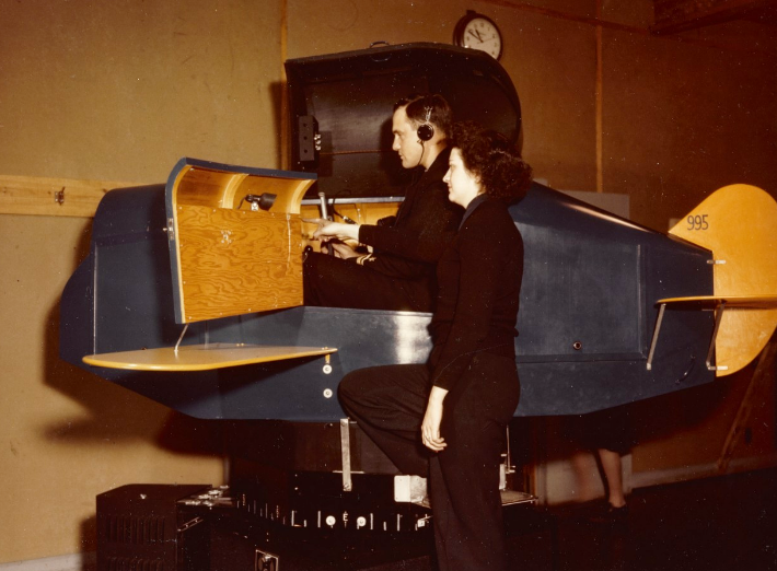 A woman stands beside a man sitting inside a helicopter replica with headphones on. The woman is pointing.